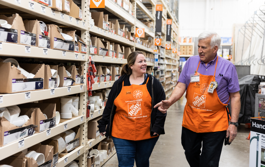 A middle-aged white woman and elderly white man in orange aprons converse in an aisle.