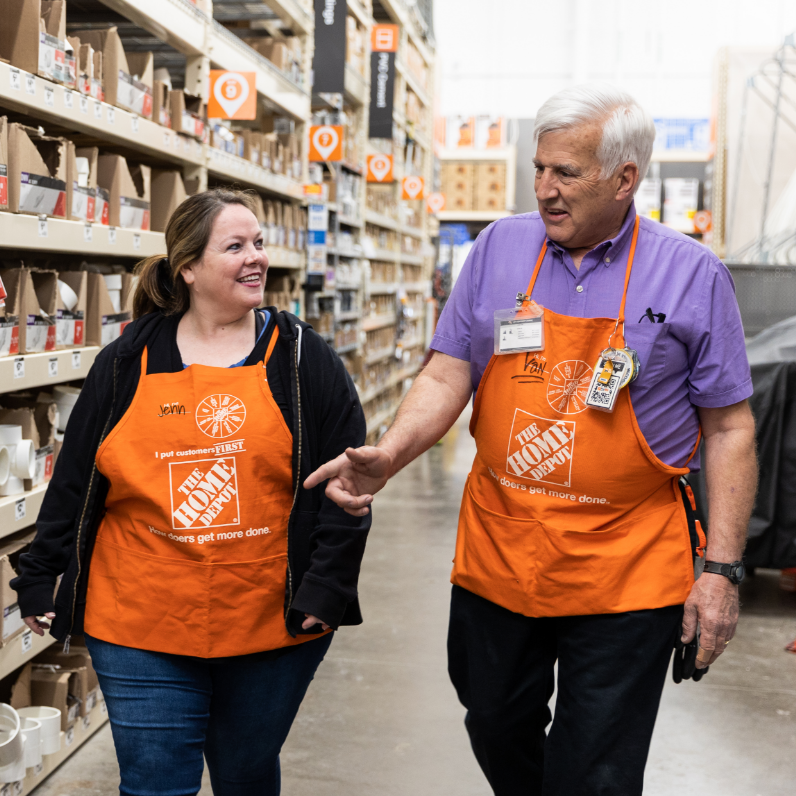 A middle-aged white woman and elderly white man in orange aprons converse in an aisle.