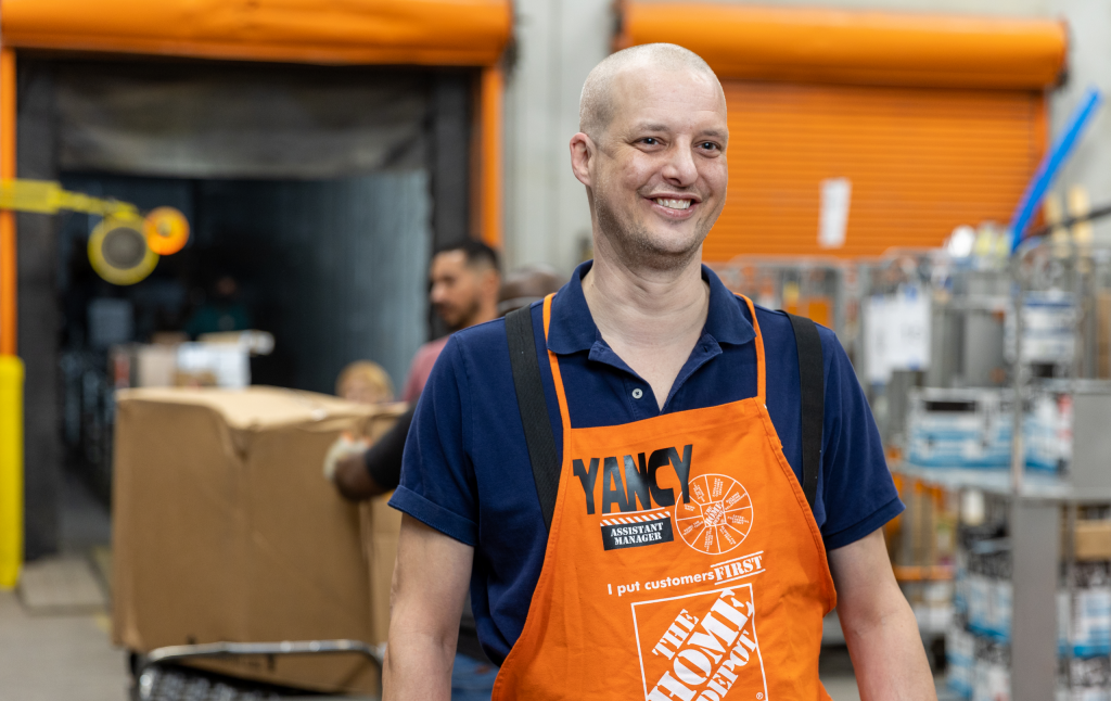 A middle-aged bald white man in an orange apron smiles while standing in a storage facility.